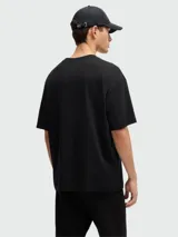 Camiseta negra de corte relajado con cuello redondo y manga corta. Presenta un diseño tridimensional en el pecho que simula un pliegue o estructura geométrica en tono negro brillante o texturizado.