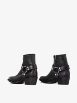 Bota corta de cuero negro con punta estilizada y taco bajo. Presenta un detalle de arnés desmontable alrededor del tobillo con argolla metálica y tachas plateadas.