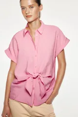 Camisa fluída color rosado confeccionada en hilatura con modal. Tiene cuello solapa, manga corta con vuelta y detalle de nudo en el delantero. Cierre frontal con botones.