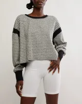 Buzo de tejido jacquard con patrón geométrico en tonos blanco roto y negro. Presenta cuello redondo, puños y bajo en rib negro, y un detalle de banda negra en las mangas.