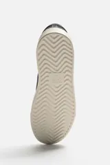 Zapatilla deportiva de hombre color gris topo, fabricada en piel con acabado serraje. Presenta cordonera elástica oculta y suela de goma de bajo volumen.
