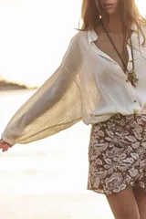 Blusa color crema de tejido arrugado con hilo metalizado, cuello redondo con escote en pico, manga larga abullonada y nudo delantero. Cierre frontal con botones con flores bordadas.