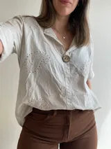 Blusa color crema con cuello camisero, mangas cortas con dobladillo y cierre frontal con un botón de madera. Presenta bordados con motivos de paisley y perforaciones.