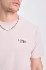 Remera de manga corta color beige con estampa en la espalda con la frase "Beach Club" y un diseño de un surfista en la playa.