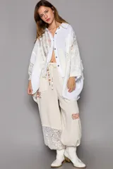 Camisa blanca de manga larga con detalles de encaje y crochet, cuello con escote en V y cierre frontal con botones. Diseño oversized con dobladillo asimétrico.