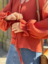 Blusa color terracota de muselina con escote en V y lazos para ajustar. Tiene mangas largas abullonadas con volados en los puños.
