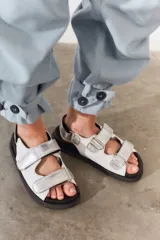 Sandalias de cuero color blanco con doble tira plateada con velcro, base negra y hebilla plateada.