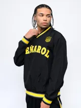 Buzo negro de nylon con cuello en V, puños y pretina con detalles en amarillo y gris. Estampado frontal con la palabra "Peñarol" en amarillo y escudo del club en la manga. Corte clásico.