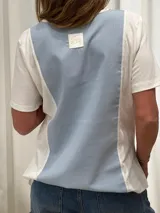 Remera blanca con panel central celeste, cuello redondo y mangas cortas. Presenta detalles de puntilla en el panel lateral.