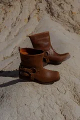 Bota corta de cuero color marrón, con taco cuadrado y punta redondeada. Presenta una correa con hebillas y argolla metálica decorativa alrededor del tobillo. Cuenta con cierre lateral.