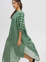 Vestido largo con mangas 3/4 y cuello mao, confeccionado en tela con estampado de círculos en tonos verde oliva y celeste pálido. Presenta cierre frontal de botones marrones y corte amplio con aberturas laterales.