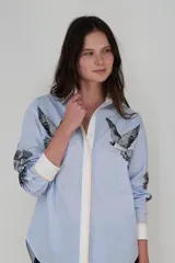 Camisa celeste a rayas verticales blancas, de corte clásico, con cuello camisero, cierre frontal con botones, mangas largas con puños abotonados y estampado de aves en el frente y las mangas.