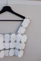 Top blanco confeccionado completamente a mano con tiras de cuero unidas por ojales metálicos, creando un efecto de cota de malla o armadura. Presenta un tirante ancho en el hombro.