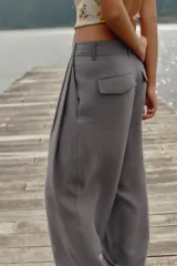 Pantalón negro de tiro alto con trabillas, doble pliegue, bolsillos laterales y tapetas en la espalda. Tiene pernera ancha y cierre frontal con cremallera y gancho metálico.