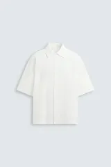 Camisa de corte relajado en color blanco, confeccionada en tejido de algodón con textura de aplicaciones. Posee cuello bowling, manga corta y cierre frontal con botones.