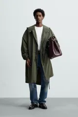 Parka verde militar de tejido técnico ligero, corte oversize, cuello alto con capucha ajustable, manga larga con puños elásticos, cintura ajustable con cordón interno, bolsillos con solapa, bajo asimétrico con abertura trasera ajustable y cierre frontal con cremallera oculta y botones a presión.