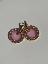 Aros colgantes con forma circular, con centro de resina color lila y borde con strass. El arito es de acero quirúrgico color dorado.