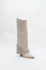 Bota texana de caña alta color beige confeccionada en gamuza, con punta fina, taco cuadrado de madera y detalle de flecos laterales. 