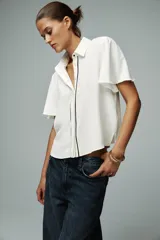 Camisa blanca de corte recto, con cuello solapa y mangas cortas. Presenta vivos negros a contraste en el frente y aberturas laterales en el ruedo. Cierre frontal con botones ocultos.