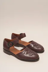 Zapato marrón de cuero con correa al tobillo y hebilla negra. Diseño trenzado en la parte delantera.