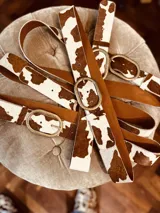 Cinturón de cuero con estampado de vaca en blanco y marrón, hebilla metálica ovalada color bronce.