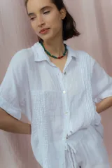 Camisa color crudo confeccionada en lino, de corte holgado, con cuello clásico, mangas cortas con dobladillo y cierre frontal con botones. Presenta detalle de pliegues verticales en la parte frontal.