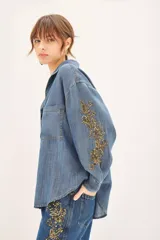 Camisa de denim azul de corte amplio, manga larga, cuello camisero y bolsillos en el delantero. Presenta bordados dorados de mostacillas, lentejuelas y canutillos en mangas y parte inferior de un lateral del pantalón.