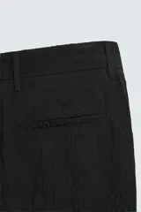 Pantalón negro de corte relajado (relaxed fit) con textura arrugada, tiro medio, bolsillos laterales y bolsillos traseros de vivo. Cierre frontal con cremallera y gancho metálico.