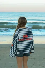 Buzo de manga larga reversible. Un lado es a rayas horizontales azul marino y blanco, con un logo rojo bordado en el pecho y un parche rojo en la manga. El otro lado es de color azul claro liso, con un cuello redondo deshilachado.