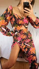 Vestido midi con estampado floral en tonos lila, amarillo y verde. Tiene escote fruncido con lazo ajustable, mangas largas abullonadas y tajo lateral.