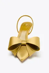Zapato de tacón destalonado color dorado, con efecto satinado, punta fina, tira trasera elástica y maxi lazo decorativo en la parte delantera.
