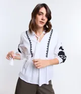 Blusa amplia de viscolino color blanco con bordados florales negros en mangas y escote, lazo en el cuello y mangas 3/4 con puño ajustado.