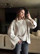 Sweater tejido color beige con cuello alto y mangas largas. Presenta aberturas verticales con detalles en marrón y negro.