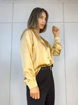 Camisa color dorado de satén plisado, con cuello clásico, cierre frontal con botones y mangas largas con puños abotonados.