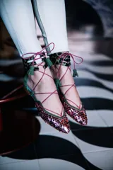 Bailarinas chatitas con punta cubierta de lentejuelas rosadas, laterales y talón de gamuza verde oliva, y tobillera de cuero con estampado animal print. Se atan al tobillo con cordones rosas y tienen un pequeño fleco decorativo en el talón.