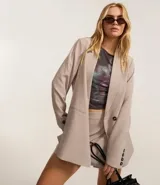 Blazer largo color beige, de corte recto, con cuello solapa, cierre frontal con doble botón, bolsillos tipo ojal y mangas largas con botones decorativos en los puños.