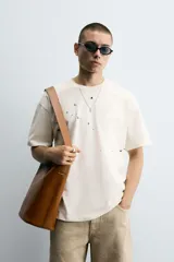 Camiseta color crudo de corte holgado, cuello redondo y manga corta. Tiene estampado de salpicaduras de pintura negra en el frente y texto estampado en la espalda.
