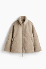 Campera de sherpa suave en color beige, de corte holgado con cuello elevado y cierre frontal bidireccional. Presenta bolsillos frontales tipo canguro y detalles de ribetes en tejido revestido.
