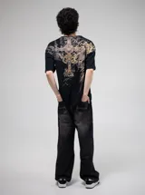 Remera negra de algodón con estampado frontal grande en tonos grises y dorados, con tipografía gótica, un diseño de cruz ornamentada y un felino en el centro.