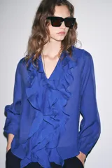 Camisa azul con escote en pico, cierre frontal con botones, mangas largas abullonadas con elástico en los puños y detalle de volantes en el frente.