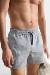 Short de baño con textura seersucker a rayas verticales azul marino y blanco. Tiene cintura elástica ajustable con cordón, bolsillos frontales y traseros, aberturas laterales en el bajo y forro interior.