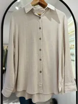Camisa beige de lino, de corte recto y manga larga con puños arremangados. Tiene cuello camisero, cierre frontal con botones de coco y aberturas laterales.