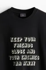 Remera negra de corte holgado con estampado frontal con la frase "Keep your friends close and your enemies far away".