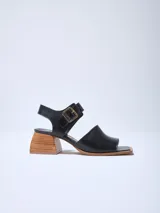 Sandalias de cuero negras con punta cuadrada, tira ancha sobre el empeine y pulsera con hebilla dorada al tobillo. Taco geométrico de madera clara.