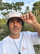 Gorro de visera curva con diseño a rayas verticales en tonos rosado pálido y blanco. La corona tiene un bordado en azul claro con texto. La visera es de color beige claro. Incluye un cordón negro ajustable para colgar del cuello.