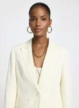 Blazer color blanco roto, de tejido de punto con corte entallado, cuello, dos bolsillos de ribete con solapa, mangas largas con puños abotonados y cierre frontal con botones.