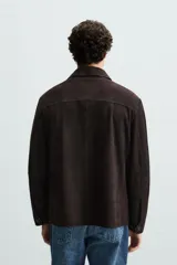 Sobrecamisa marrón de cuero gamuzado, corte recto, con cuello clásico, cierre frontal con botones, mangas largas con puños abotonados y bolsillos de parche en el pecho.