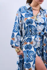 Vestido estilo camisola con estampado floral en tonos azules y marrones, con cuello camisero, mangas largas con puños y cierre frontal con botones. Incluye cinturón de la misma tela.