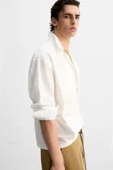 Camisa blanca de corte relaxed fit confeccionada en tejido de algodón y lino. Tiene cuello bowling, manga larga con puños abotonados, jaretas en el delantero, bolsillos de parche en pecho y cadera, bajo con aberturas laterales y cierre frontal con botones.