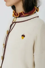 Cárdigan tejido en 50% algodón y 50% acrílico, color crema, con pespuntes tejidos en contraste color borgoña y bordado de limón en el pecho. Tiene cuello con volados, cierre frontal con botones y corte relajado.